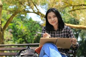 sorridente attraente asiatico giovane donna scrittura per fare elenco e organizzazione sua lavoro su taccuino nel città parco foto