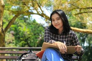 sorridente attraente asiatico giovane donna scrittura per fare elenco e organizzazione sua lavoro su taccuino nel città parco foto