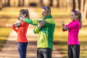 tre dilettante atleti maturo uomo e Due giovane donne siamo riscaldamento su prima jogging, fare superiore corpo allungamento nel il parco foto