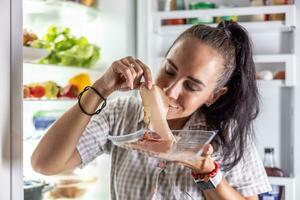 molto Affamato donna nel pigiama godendo prosciutto a notte di il frigo foto