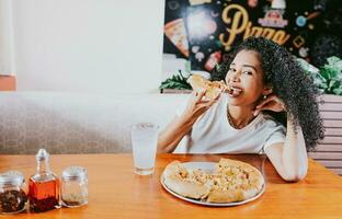 sorridente giovane donna godendo un' Pizza nel un' ristorante. contento afro capelli ragazza mangiare Pizza nel un' ristorante, mangiare e guardare a telecamera foto