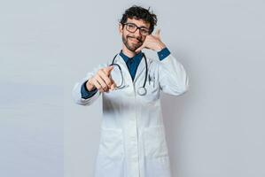 sorridente medico fabbricazione chiamata gesto e puntamento a il telecamera. giovane medico fabbricazione chiamata gesto isolato foto