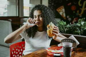 ritratto di un afro ragazza godendo Hamburger nel un' ristorante. latino donna suzione sua dita Tenere un' Hamburger nel un' ristorante foto