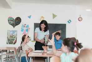 femmina insegnante con bambini nel biologia classe a elementare scuola conduzione biologia o botanico scientifico sperimentare di sostenibile in crescita impianti. apprendimento di impianti nel un' bicchiere vaso foto