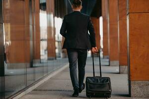 andando per aeroporto terminale. fiducioso uomo d'affari viaggiatore a piedi su città strade e traino il suo valigia potabile caffè e A proposito di su smartphone foto