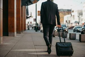 andando per aeroporto terminale. fiducioso uomo d'affari viaggiatore a piedi su città strade e traino il suo valigia potabile caffè e A proposito di su smartphone foto