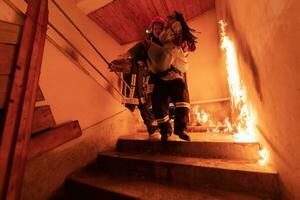 coraggioso vigile del fuoco discende le scale di un' ardente edificio e detiene salvato ragazza nel il suo braccia. Aperto fuoco e uno pompiere nel il sfondo. foto