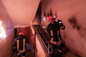 coraggioso vigile del fuoco discende le scale di un' ardente edificio e detiene salvato ragazza nel il suo braccia. Aperto fuoco e uno pompiere nel il sfondo. foto