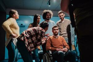colleghi Aiuto loro amico nel un' sedia a rotelle per prendere il suo primo passaggi nel un' moderno ufficio dopo un' grave malattia foto