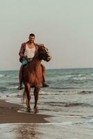 un' moderno uomo nel estate Abiti gode equitazione un' cavallo su un' bellissimo sabbioso spiaggia a tramonto. selettivo messa a fuoco foto