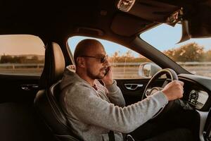 un' uomo con un' occhiali da sole guida un' auto e parlando su smartphone a tramonto. il concetto di auto viaggio foto