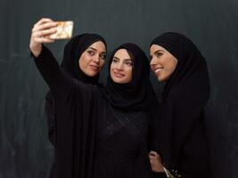 gruppo di giovane bellissimo musulmano donne nel alla moda vestito con hijab utilizzando smartphone mentre assunzione autoscatto immagine nel davanti di nero sfondo foto