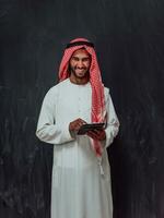 giovane arabo musulmano uomo d'affari utilizzando tavoletta computer indossare hijab Abiti nel davanti di nero lavagna foto
