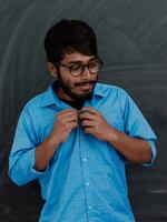 indiano giovane alunno indossare bicchieri mette su un' blu camicia mentre in piedi nel davanti di il scuola lavagna foto