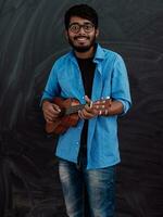 indiano giovane uomo nel un' blu camicia e bicchieri giocando il chitarra nel davanti di il scuola lavagna foto