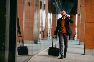 andando per aeroporto terminale. fiducioso uomo d'affari viaggiatore a piedi su città strade e traino il suo valigia potabile caffè e A proposito di su smartphone foto