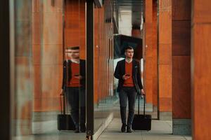 andando per aeroporto terminale. fiducioso uomo d'affari viaggiatore a piedi su città strade e traino il suo valigia potabile caffè e A proposito di su smartphone foto