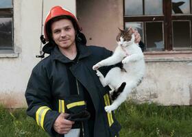 avvicinamento ritratto di eroico vigile del fuoco nel protettivo completo da uomo e rosso casco detiene salvato gatto nel il suo braccia. pompiere nel fuoco combattente operazione. foto