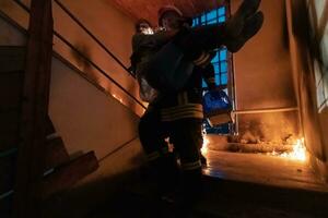 coraggioso vigile del fuoco discende le scale di un' ardente edificio e detiene salvato ragazza nel il suo braccia. Aperto fuoco e uno pompiere nel il sfondo. foto