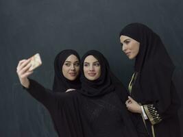 gruppo di giovane musulmano donne nel alla moda vestito con hijab utilizzando smartphone mentre assunzione autoscatto immagine nel davanti di nero sfondo foto