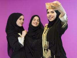 gruppo di bellissimo musulmano donne Due di loro nel alla moda vestito con hijab utilizzando mobile Telefono mentre assunzione autoscatto immagine isolato su rosa sfondo che rappresentano moderno Islam moda tecnologia foto