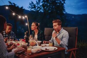 un' gruppo di giovane diverso persone avendo cena su il terrazza di un' moderno Casa nel il sera. divertimento per amici e famiglia. celebrazione di vacanze, matrimoni con barbecue. foto