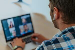 il uomo seduta a un' tavolo nel un' moderno vivente camera, con cuffie utilizzando un' il computer portatile per attività commerciale video Chiacchierare, conversazione con amici e divertimento foto