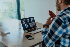 il uomo seduta a un' tavolo nel un' moderno vivente camera, con cuffie utilizzando un' il computer portatile per attività commerciale video Chiacchierare, conversazione con amici e divertimento foto