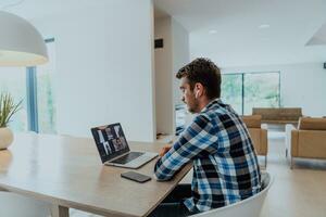 il uomo seduta a un' tavolo nel un' moderno vivente camera, con cuffie utilizzando un' il computer portatile per attività commerciale video Chiacchierare, conversazione con amici e divertimento foto