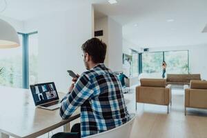 il uomo seduta a un' tavolo nel un' moderno vivente camera, utilizzando un' smartphone e il computer portatile per attività commerciale video Chiacchierare, conversazione con amici e divertimento foto