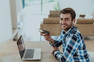 il uomo seduta a un' tavolo nel un' moderno vivente camera, con cuffie utilizzando un' il computer portatile e smartphone per attività commerciale video Chiacchierare, conversazione con amici e divertimento foto