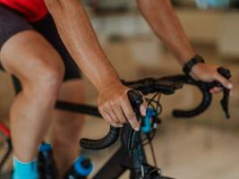 avvicinamento foto di uomo equitazione un' Triathlon bicicletta su un' macchina simulazione nel un' moderno vivente camera. formazione durante pandemia condizioni.