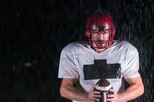 americano calcio campo solitario atleta guerriero in piedi su un' campo detiene il suo casco e pronto per giocare a. giocatore preparazione per correre, attacco e Punto touchdown. piovoso notte con drammatico nebbia, blu leggero foto