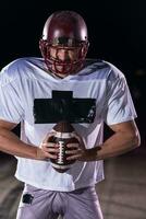 americano calcio campo solitario atleta guerriero in piedi su un' campo detiene il suo casco e pronto per giocare a. giocatore preparazione per correre, attacco e Punto touchdown. piovoso notte con drammatico nebbia, blu leggero foto