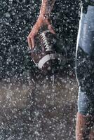 vicino su di americano calcio atleta guerriero in piedi su un' campo messa a fuoco su palla e pronto per giocare a. giocatore preparazione per correre, attacco e Punto touchdown. piovoso notte con drammatico lente bagliore e pioggia gocce. foto