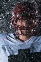 americano calcio campo solitario atleta guerriero in piedi su un' campo detiene il suo casco e pronto per giocare a. giocatore preparazione per correre, attacco e Punto touchdown. piovoso notte con drammatico nebbia, blu leggero foto