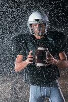 americano calcio campo solitario atleta guerriero in piedi su un' campo detiene il suo casco e pronto per giocare a. giocatore preparazione per correre, attacco e Punto touchdown. piovoso notte con drammatico nebbia, blu leggero foto