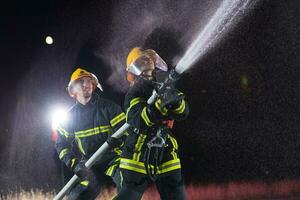 i vigili del fuoco utilizzando un' acqua tubo flessibile per eliminare un' fuoco rischio. squadra di femmina e maschio pompiere nel pericoloso salvare missione. foto