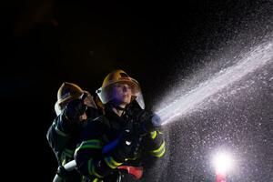 i vigili del fuoco utilizzando un' acqua tubo flessibile per eliminare un' fuoco rischio. squadra di femmina e maschio pompiere nel pericoloso salvare missione. foto