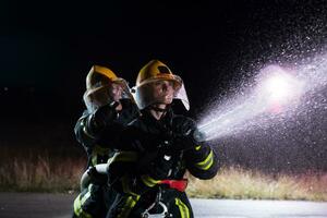 i vigili del fuoco utilizzando un' acqua tubo flessibile per eliminare un' fuoco rischio. squadra di femmina e maschio pompiere nel pericoloso salvare missione. foto