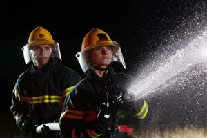 i vigili del fuoco utilizzando un' acqua tubo flessibile per eliminare un' fuoco rischio. squadra di femmina e maschio pompiere nel pericoloso salvare missione. foto