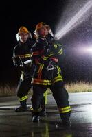 i vigili del fuoco utilizzando un' acqua tubo flessibile per eliminare un' fuoco rischio. squadra di femmina e maschio pompiere nel pericoloso salvare missione. foto