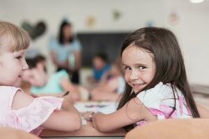 poco ragazze seduta nel elementare scuola disegno su carta con loro amici mentre seduta nel un' moderno aula foto