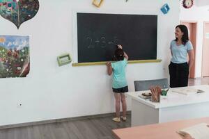 elementare scuola. il femmina insegnante porzione il bambino alunno mentre scrittura il risposta su il lavagna. foto