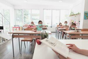 multirazziale gruppo di bambini indossare viso maschere Lavorando a classe, scrittura e ascoltando spiegazioni di insegnante nel aula foto