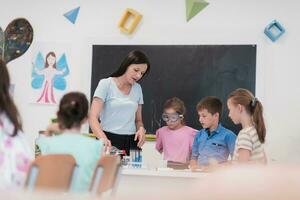 elementare scuola scienza aula entusiasta insegnante spiega chimica per diverso gruppo di figli, poco ragazzo miscele sostanze chimiche nel bicchieri. bambini imparare con interesse foto