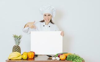 capocuoco ragazza con verdure a tavolo Tenere vuoto menù. bellissimo donna capocuoco con tavolo di verdure Tenere un' vuoto asse, ragazza capocuoco nel il cucina mostrando un' vuoto tavola foto