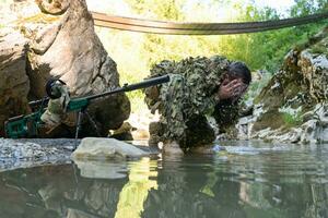 soldato nel un' camuffare completo da uomo uniforme potabile fresco acqua a partire dal il fiume. militare cecchino fucile su il lato. foto