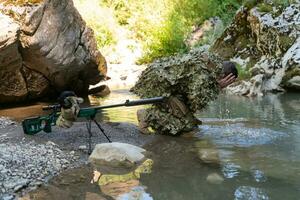 soldato nel un' camuffare completo da uomo uniforme potabile fresco acqua a partire dal il fiume. militare cecchino fucile su il lato. foto