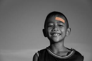 contento ragazzo Sorridi e affascinante avere plastica bendare su testa ferita, bambini incidenti e primo aiuto, bambini con cerotto, fisico ferita, stile di vita, felice incidente, nero e bianca immagine,cielo sfondo. foto